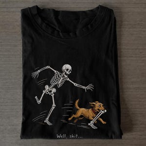 Può includere: Una t-shirt nera con la grafica di uno scheletro bianco che insegue un cane marrone che scappa con un osso in bocca. Sotto l'illustrazione, il testo "Well, shit..." è stampato in un piccolo carattere bianco senza grazie.