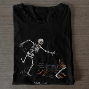 Può includere: Una t-shirt nera con una stampa grafica di uno scheletro umano che insegue un cane Doberman nero e focato che scappa con un osso in bocca. Sotto l'illustrazione, il testo "Well, shit..." è stampato in un piccolo carattere di colore bianco.