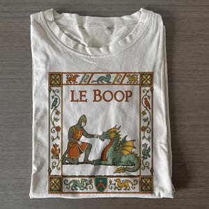 Könnte beinhalten: Ein weißes T-Shirt mit einem Grafikdruck im mittelalterlichen Stil. Das Motiv zeigt einen Ritter in Kettenrüstung, der die Nase eines grünen Drachen berührt, mit dem Text LE BOOP darüber. Die Szene ist von einem floralen Rahmen umgeben.
