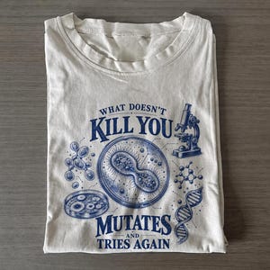 Può includere: Una t-shirt beige chiaro con una stampa grafica blu scuro raffigurante un microscopio, una piastra di Petri, un filamento di DNA e strutture cellulari. Il testo recita: WHAT DOESN'T KILL YOU MUTATES AND TRIES AGAIN. Il disegno è centrato.