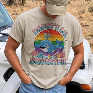 Könnte beinhalten: Ein beiges T-Shirt mit einer Grafik eines blauen Wals, der vor einem Regenbogenkreis Wasser spritzt. Der Text DADDY'S LIL SQUIRTER ist in geschwungener Schrift gedruckt. Das Design enthält Muscheln, einen Seestern und Blasen als Illustration.