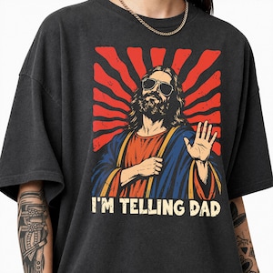 May include: A charcoal gray short-sleeve t-shirt featuring a graphic of a bearded figure wearing sunglasses, dressed in a blue and orange robe, set against a red sunburst background with the text I'M TELLING DAD printed in cream-colored letters.