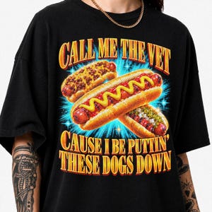 Peut inclure: Un t-shirt noir avec une impression graphique de trois hot-dogs garnis. Le texte CALL ME THE VET CAUSE I BE PUTTIN' THESE DOGS DOWN est écrit en lettres jaunes et orange sur un fond bleu vif avec un effet d'éclat d'étoile.