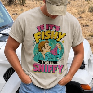 Könnte beinhalten: Ein beiges Kurzarm-T-Shirt mit einer grafischen Illustration einer Person mit Schnurrbart, die eine rosa Unterwäsche mit Fischmuster hochhält. Der Text IF IT'S FISHY I WILL SNIFFY steht in gelben und pinken Buchstaben auf einem türkisen Kreis.