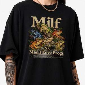 Könnte beinhalten: Ein schwarzes T-Shirt mit einem Grafikdruck verschiedener bunter Frösche auf Baumstämmen und Laub. Der Text lautet Milf Man I Love Frogs, gefolgt von einem kleinen beschreibenden Absatz über die Ordnung Anura in beiger Vintage-Schriftart.