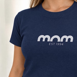 Può includere: Una t-shirt blu navy a maniche corte con la parola "mom" in un carattere bianco, grassetto e arrotondato sul petto. Sotto la parola "mom", il testo "EST 1994" è stampato in un carattere bianco più piccolo e sottile.