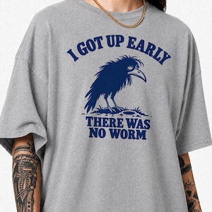 Könnte beinhalten: Ein graues T-Shirt mit einem dunkelblauen Aufdruck eines zerzausten Raben, der über einem Loch im Boden steht. Der Text über dem Raben lautet I GOT UP EARLY und der Text unter dem Raben lautet THERE WAS NO WORM in einer fetten blauen Schrift.