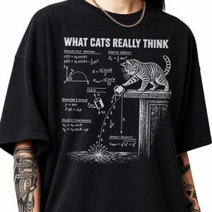 Pode incluir: Uma t-shirt preta com uma estampa gráfica branca intitulada WHAT CATS REALLY THINK. O design mostra um gato a empurrar uma caneca de uma borda, acompanhado por equações físicas de movimento, energia cinética e velocidade.