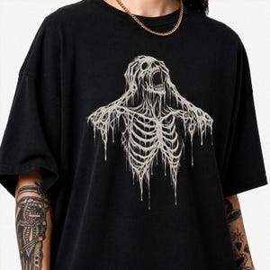Può includere: Una t-shirt nera a maniche corte con una stampa grafica di uno scheletro con le mani sulla testa, la bocca aperta in un grido e dettagli gocciolanti e sciolti di colore bianco sporco sulla parte del petto dell'indumento.