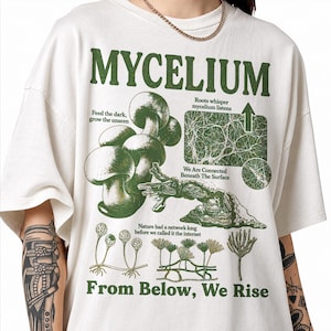 Op de afbeelding: Een gebroken wit t-shirt met een groene grafische print met de tekst MYCELIUM, Feed the dark, grow the unseen, Roots whisper mycelium listens, We Are Connected Beneath The Surface, Nature had a network long before we called it the internet, en From Below, We Rise, samen met illustraties van paddenstoelen en wortelsystemen.