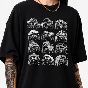 May include: A black short-sleeved t-shirt featuring a white graphic print of twelve distinct cat faces. Each cat is depicted wearing a different accessory, including hats, a crown, a masquerade mask, and a beret, arranged in a grid pattern.