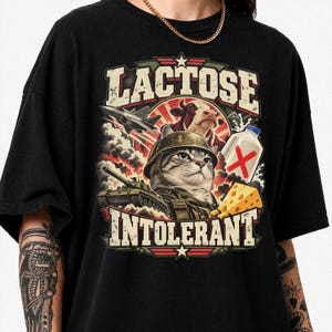 Könnte beinhalten: Ein schwarzes T-Shirt mit einem Grafikdruck, der eine Katze mit Militärhelm, einen Panzer, einen Kampfjet, eine Kuh, einen Milchkarton mit einem roten X und ein Stück Käse zeigt. Der Text LACTOSE INTOLERANT ist in großen, fetten Buchstaben gedruckt.