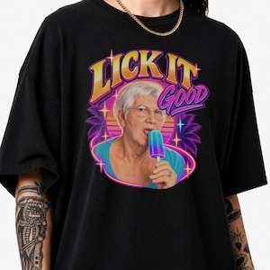 Op de afbeelding: Een zwart t-shirt met een grafische print van een ouder persoon met kort wit haar en een bril die aan een blauw en paars ijsje likt. De tekst LICK IT GOOD staat in geel en paars boven de persoon, omlijst door palmbomen en sterren.