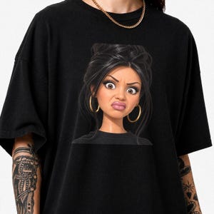 Könnte beinhalten: Ein schwarzes T-Shirt mit kurzen Ärmeln und einer zentralen grafischen Illustration einer Zeichentrickfigur mit dunklem Haar, großen Augen und goldenen Creolen. Die Figur hat einen skeptischen Gesichtsausdruck mit geschürzten Lippen.