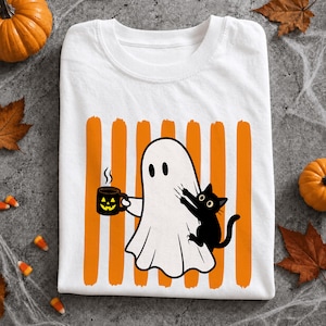 Può includere: Una t-shirt bianca con la grafica di un fantasma bianco che tiene in mano una tazza nera con una faccia di zucca gialla e vapore, mentre un gatto nero si aggrappa al fantasma, il tutto su uno sfondo a strisce verticali arancioni.