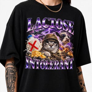 Op de afbeelding: Een zwart t-shirt met korte mouwen en een grafische print van een kat met een militaire helm, een koe met een zonnebril, een melkfles met een rode X, kaasblokjes, gevechtsvliegtuigen en een explosie, met de tekst LACTOSE INTOLERANT in het paars.