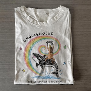May include: A cream-colored t-shirt featuring a graphic of an orca whale with a brown creature wearing a party hat and holding a toy gun on its back. A rainbow swirl, stars, and the text "UNDIAGNOSED but something ain't right" are printed on the front.
