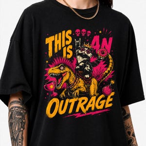 Peut inclure: Un t-shirt noir avec une impression graphique représentant un chat portant une couronne et des lunettes en forme d'étoile chevauchant un T-Rex à crête rose. Le motif inclut un trophée, un donut, trois crânes, une bombe et le texte THIS IS AN OUTRAGE en jaune.