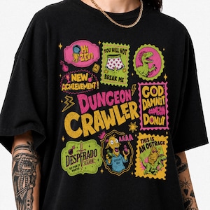 Könnte beinhalten: Ein schwarzes T-Shirt mit einem bunten Grafikdruck und dem Text "DUNGEON CRAWLER" in großen gelben Buchstaben. Das Design enthält verschiedene kleinere rechteckige und ovale Rahmen mit Illustrationen eines Dinosauriers, einer Ziege und Text.