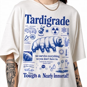 Può includere: Una t-shirt a maniche corte color bianco sporco con una stampa grafica blu di un tardigrado. Il design include icone scientifiche e il testo "Tardigrade", "we survive everything so you don't have to" e "Tiny, Tough & Nearly Immortal!" insieme ad altre scritte.