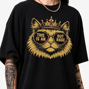 Pode incluir: Uma t-shirt preta com o gráfico de um gato dourado a usar uma coroa com uma gema vermelha e óculos redondos. O texto "THIS IS AN" está impresso na lente esquerda e "OUT RAGE" na lente direita dos óculos do gato.