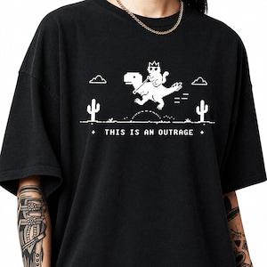 Può includere: Una t-shirt nera con una grafica in pixel art bianca che raffigura un gatto con una corona in sella a un dinosauro. La scena include cactus, nuvole e il testo "THIS IS AN OUTRAGE" stampato in un font pixelato sotto l'illustrazione.