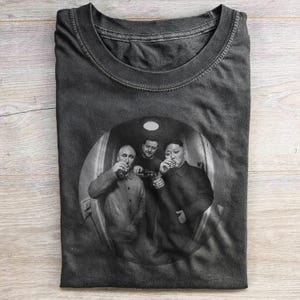 Könnte beinhalten: Ein anthrazitfarbenes T-Shirt mit Rundhalsausschnitt und einem kreisförmigen schwarz-weißen Grafikdruck von drei Personen in Mänteln, die kleine Gläser halten, aufgenommen mit einem Fischaugenobjektiv-Effekt, mittig auf der Brust platziert.