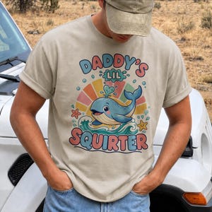 Könnte beinhalten: Ein beiges T-Shirt mit einer Grafik eines blauen Wals, der Wasser spritzt, umgeben von einem bunten Sonnenstrahlenmuster, Wellen und Seesternen. Der Text "DADDY'S LIL SQUIRTER" ist in einer fetten, mehrfarbigen Schrift über und unter dem Wal gedruckt.