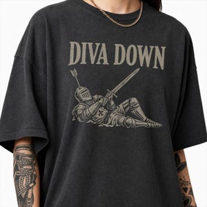 May include: A charcoal grey t-shirt featuring a graphic of a knight in armour lying down, holding a sword with an arrow protruding from the helmet. The text DIVA DOWN is printed in a light beige colour above the knight illustration on the chest.