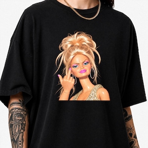 Könnte beinhalten: Ein schwarzes T-Shirt mit kurzen Ärmeln und einem Grafikdruck einer blonden Puppe mit hohem, unordentlichem Dutt, die goldene Creolen und ein goldenes Paillettenoberteil trägt und mit einer pink lackierten Hand den Mittelfinger zeigt.