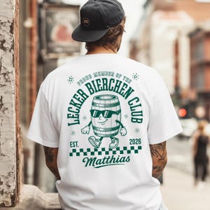 Puede incluir: Una camiseta blanca con un gráfico verde en la espalda. El diseño muestra un barril de cerveza de dibujos animados con gafas de sol y zapatos, rodeado por el texto "PROUD MEMBER OF THE LECKER BIERCHEN CLUB", "EST. 2026" y el nombre "Matthias".