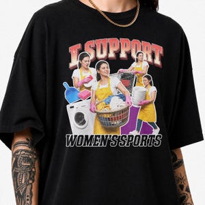 May include: A black t-shirt featuring a graphic print with the text I SUPPORT WOMEN'S SPORTS. The design shows multiple images of a person wearing a yellow apron and pink gloves performing household chores like laundry, cleaning, and shopping.