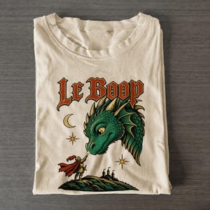Könnte beinhalten: Ein beiges T-Shirt mit einem Grafikdruck eines grünen Drachenkopfes, eines Ritters mit rotem Umhang, einer Mondsichel und Sternen. Der Text "Le Boop" ist in einer fetten, stilisierten roten Schrift über dem Drachen abgebildet.