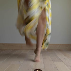 May include: A small amber glass candle jar with a lit wick and a beige label featuring the text GRUMPY X SUNSHINE. The candle sits on a light wood floor, while a blurred figure in a yellow and white patterned garment moves in the background.
