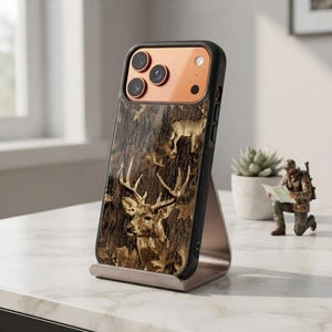 May include: A smartphone case featuring a camouflage pattern with deer imagery, including a prominent buck with antlers. The case has a black frame and a copper-coloured camera module area. It is displayed upright on a metallic stand.