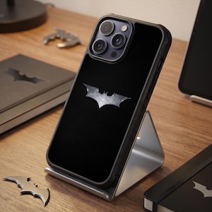Könnte beinhalten: Eine schwarze Smartphone-Hülle mit einem metallisch silbernen Batman-Logo auf der Rückseite. Die Hülle hat eine strukturierte Oberfläche und steht auf einem silbernen Metallständer. Das Telefon verfügt über ein Dreifach-Kamerasystem mit Blitz.