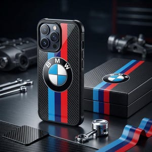 Op de afbeelding: Een zwarte smartphonehoes met koolstofvezeltextuur, voorzien van het BMW-logo en verticale blauwe, donkerblauwe en rode strepen. De hoes heeft een uitsparing voor een drievoudig camerasysteem en wordt getoond met een bijpassende doos en zuiger.