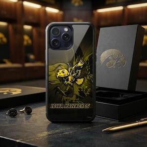 Puede incluir: Una funda de smartphone negra con un gráfico amarillo y negro de una mascota halcón y el texto IOWA HAWKEYES. La funda se muestra junto a una caja de regalo negra con un logotipo de halcón dorado, dos gemelos negros y un bolígrafo dorado.