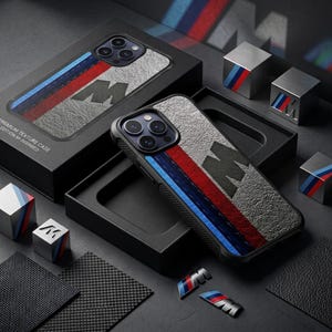 Op de afbeelding: Een zwarte smartphonehoes met een getextureerd grijs oppervlak, voorzien van een verticaal streeppatroon in blauw, donkerblauw en rood. De hoes toont een groot zwart M-logo. Het wordt gepresenteerd in een zwarte doos met tekst PREMIUM TEXTURE CASE.