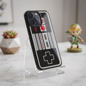 May include: A black smartphone case featuring a graphic design inspired by a vintage game controller, including red buttons, grey directional pads, and the text Nintendo, Select, and Start printed on the back surface of the protective cover.