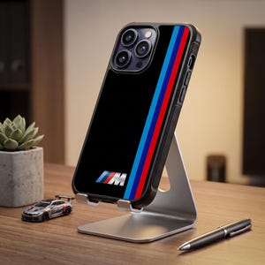 May include: A black smartphone case featuring the M logo and a vertical stripe pattern in light blue, dark blue, and red. The case has a textured side grip and is mounted on a silver metal stand, displaying a triple-lens camera array on the back.