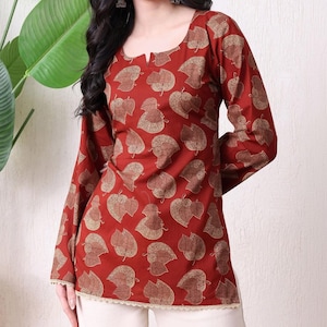 May include: A long-sleeved tunic in a deep rust-red colour featuring a repeating gold-toned leaf pattern. The garment has a scoop neckline with a small central slit and a delicate cream-coloured lace trim along the bottom hemline.