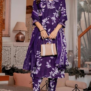 Pu&ograve; includere: Un completo kurta viola scuro con motivo a foglie bianche, scollo a V con decorazioni argentate e maniche a tre quarti. L'outfit include pantaloni dritti abbinati e un dupatta coordinato, completato da una borsa rettangolare beige.