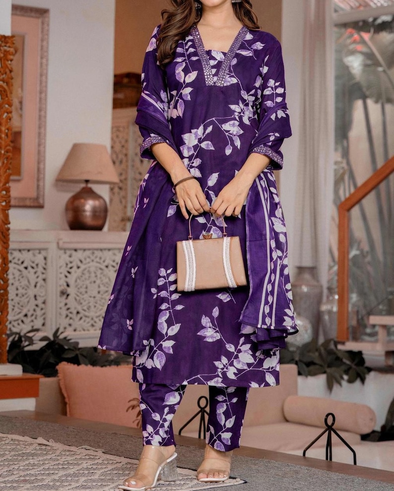 Pu&ograve; includere: Un completo kurta viola scuro con motivo a foglie bianche, scollo a V con decorazioni argentate e maniche a tre quarti. L'outfit include pantaloni dritti abbinati e un dupatta coordinato, completato da una borsa rettangolare beige.
