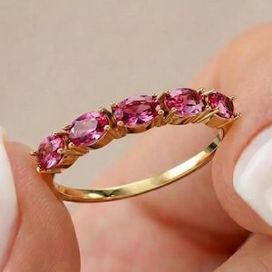 May include: A gold band ring featuring five oval-cut pink gemstones arranged in a row. The stones are set in a prong setting along the top half of the thin, polished gold band, creating a continuous line of vibrant pink colour across the finger.