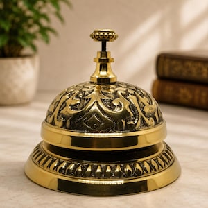 Puede incluir: Un timbre de escritorio de latón pulido con un patrón floral en relieve en su cúpula. El timbre tiene una base circular lisa con un borde decorativo y un pequeño pomo con cuentas en la parte superior para su activación manual, acabado metálico.