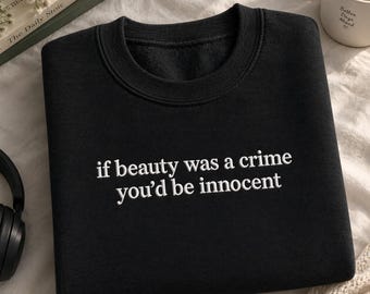 May include: A black crewneck sweatshirt featuring white embroidered text centred on the chest that reads, if beauty was a crime you’d be innocent. The garment has a ribbed collar, cuffs, and hem, with a soft fleece interior visible at the neckline.