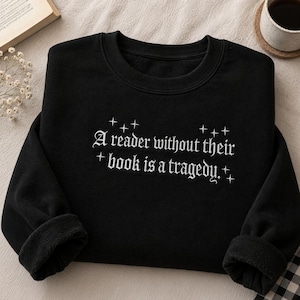 May include: A black long-sleeve sweatshirt featuring white embroidered text that reads, A reader without their book is a tragedy. The text is surrounded by small, four-pointed star icons, and the garment has a ribbed crew neckline and cuffs.