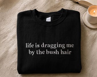 May include: A black crewneck sweatshirt featuring white embroidered text on the chest that reads, life is dragging me by the bush hair. The garment has long sleeves, a ribbed neckline, and a standard fit, presented in a clean, folded layout.