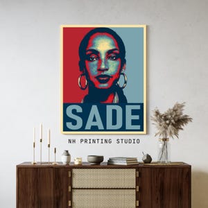 May include: A rectangular wall art print featuring a stylised portrait of Sade in red, blue, and black tones. The name SADE is printed in large, bold, light blue capital letters at the bottom. Below the print, the text NH PRINTING STUDIO is visible.